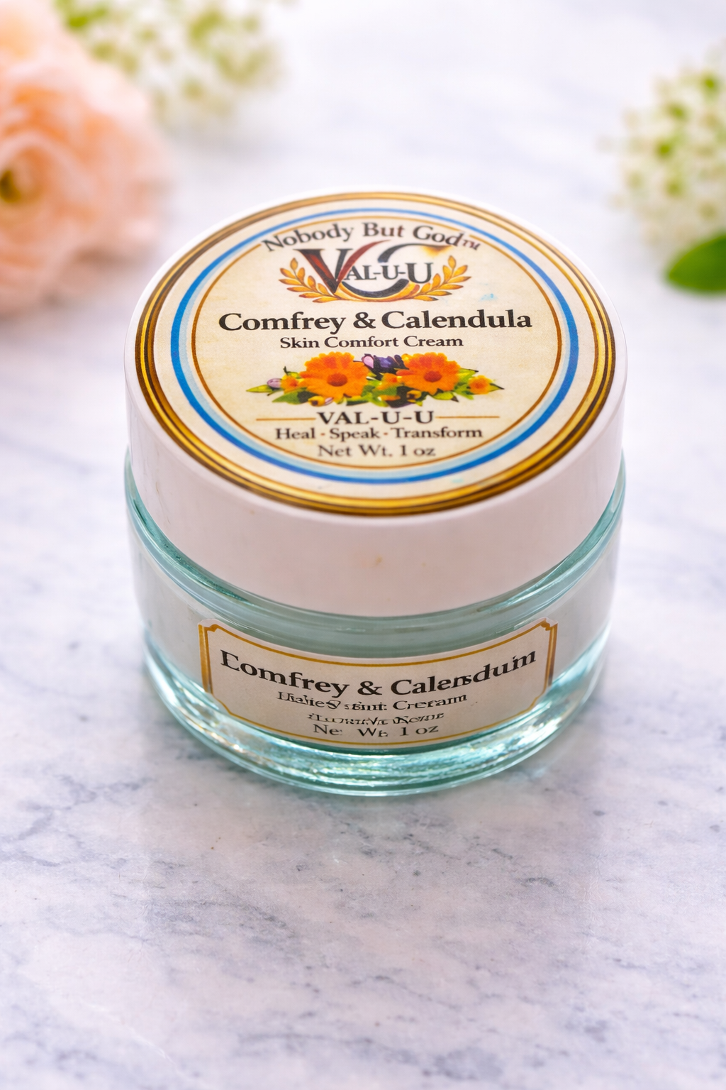 Jar of Val-U-U Comfrey & Calendula Skin Comfort Cream on a marble surface with flowers in the background